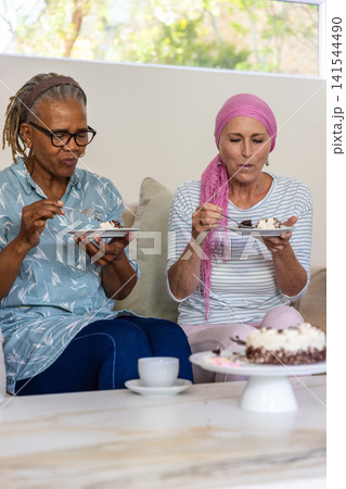 Diverse senior friends sitting on couch in living room holding plates with chocolate cake slices Diverse senior friends sitting on couch in living room holding plates with chocolate cake slices 141544490