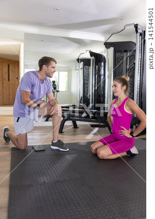 Male instructor and client kneeling in gym on black mat planning workout with tablet, machine Male instructor and client kneeling in gym on black mat planning workout with tablet, machine 141544501