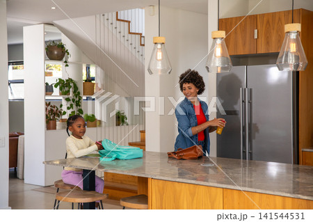 Diverse mother and daughter unpacking turquoise backpack on kitchen island with glass of juice Diverse mother and daughter unpacking turquoise backpack on kitchen island with glass of juice 141544531