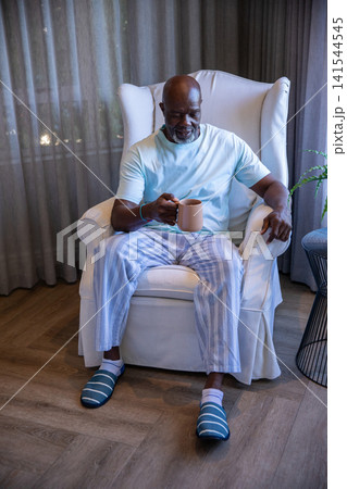 Senior African American man sitting in armchair in living room wearing slippers, holding mug Senior African American man sitting in armchair in living room wearing slippers, holding mug 141544545
