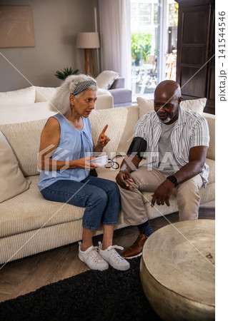 Diverse senior couple checking blood pressure with digital cuff monitor on sofa in living room 141544546