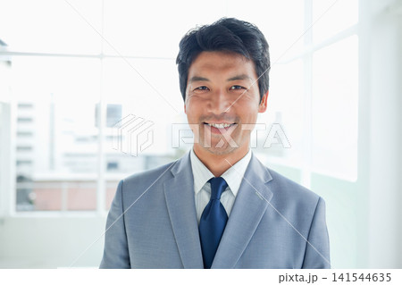 Mid-adult Asian man standing smiling in office wearing gray suit jacket navy-tie by windows Mid-adult Asian man standing smiling in office wearing gray suit jacket navy-tie by windows 141544635