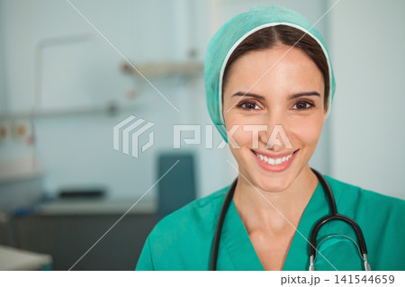 Female nurse smiling toward camera in clinic wearing green scrubs and surgical cap with stethoscope Female nurse smiling toward camera in clinic wearing green scrubs and surgical cap with stethoscope 141544659