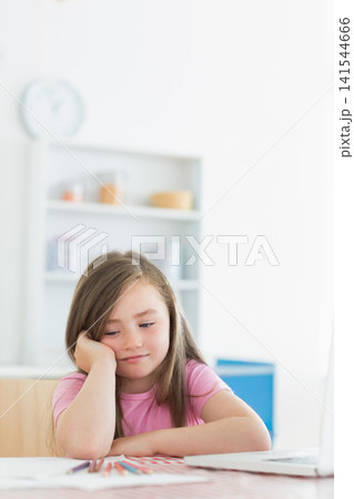 Female child leaning head on hand at table wearing pink shirt, drawing with colored pencils, laptop Female child leaning head on hand at table wearing pink shirt, drawing with colored pencils, laptop 141544666