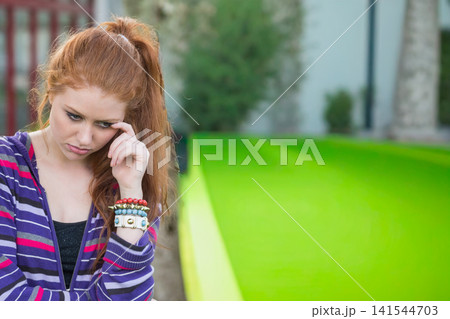 Female in 20s standing left on lawn rubbing temple, wearing purple hoodie and bracelets, copy space 141544703