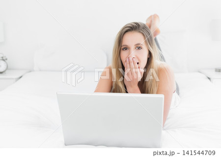 Woman 20s wearing white top lying on bed using silver laptop, covering mouth, alarm clock lamp Woman 20s wearing white top lying on bed using silver laptop, covering mouth, alarm clock lamp 141544709