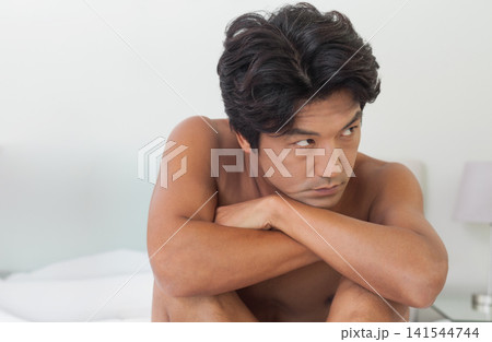 Mid adult Asian man sitting shirtless on bed, leaning forward and crossing arms, lamp in bedroom Mid adult Asian man sitting shirtless on bed, leaning forward and crossing arms, lamp in bedroom 141544744