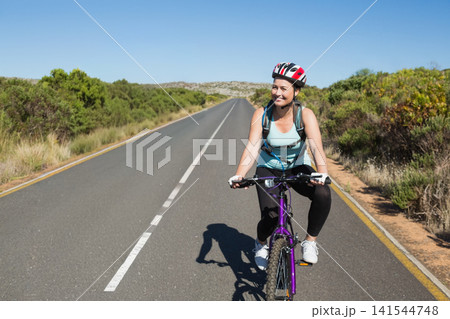 Female cyclist riding purple mountain-bike wearing helmet, gloves and pack on road with dashed-line 141544748