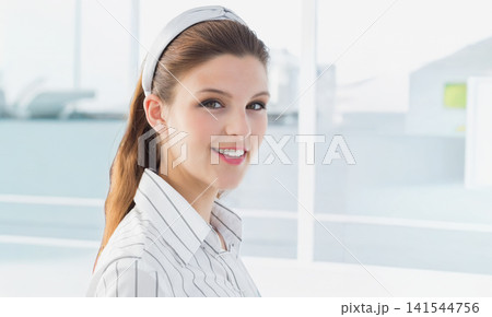 Woman 20s wearing striped shirt, headband smiling at camera at office windows with blurred rooftops Woman 20s wearing striped shirt, headband smiling at camera at office windows with blurred rooftops 141544756