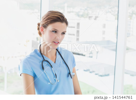 Female nurse in twenties standing in clinic near window wearing light blue scrubs stethoscope badge Female nurse in twenties standing in clinic near window wearing light blue scrubs stethoscope badge 141544761