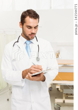 Male physician writing on clipboard by patient bed and curtain, wearing lab coat, stethoscope Male physician writing on clipboard by patient bed and curtain, wearing lab coat, stethoscope 141544771