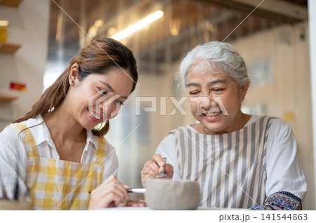 Asian Senior Woman and Adult Daughter Smiling While Making Clay Pottery in ceramic workshop Asian Senior Woman and Adult Daughter Smiling While Making Clay Pottery in ceramic workshop 141544863