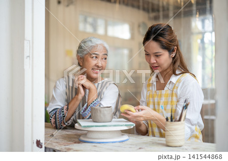 Young Asian Woman Explaining Pottery Sponge Technique to Happy Senior Mother in ceramic workshop 141544868