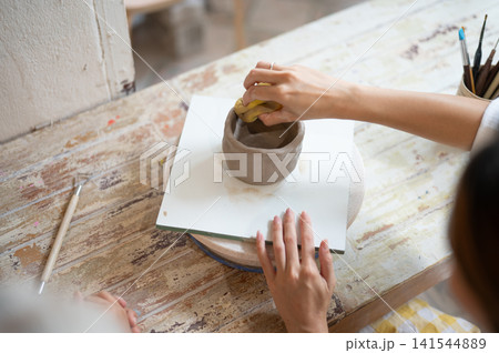 Overhead Angle of Woman's Hands Shaping a Clay Bowl with Sponge on Turntable in pottery workshop Overhead Angle of Woman's Hands Shaping a Clay Bowl with Sponge on Turntable in pottery workshop 141544889