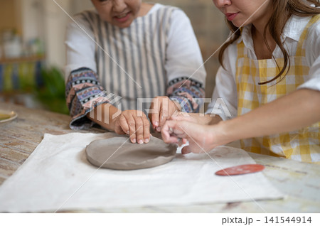 Close up of Asian Mother and Daughter Shaping Ceramic Plate Rim Together in pottery workshop Close up of Asian Mother and Daughter Shaping Ceramic Plate Rim Together in pottery workshop 141544914