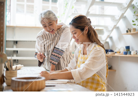 Senior Asian Woman Guiding Daughter in Clay Crafting at Studio Workbench in pottery workshop. Senior Asian Woman Guiding Daughter in Clay Crafting at Studio Workbench in pottery workshop. 141544923