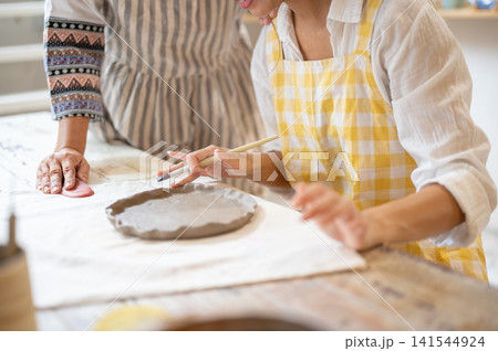 Close Up of Young Woman Painting Clay Plate with Senior Guide Beside in pottery workshop studio. Close Up of Young Woman Painting Clay Plate with Senior Guide Beside in pottery workshop studio. 141544924