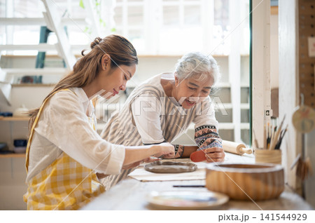 Young Asian Woman Shaping Clay Plate with Senior Mother Guiding in pottery workshop studio. Young Asian Woman Shaping Clay Plate with Senior Mother Guiding in pottery workshop studio. 141544929