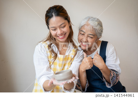 Asian Mother and Daughter Smiling and Giving Thumbs Up with Clay Bowl in pottery workshop studio. Asian Mother and Daughter Smiling and Giving Thumbs Up with Clay Bowl in pottery workshop studio. 141544961