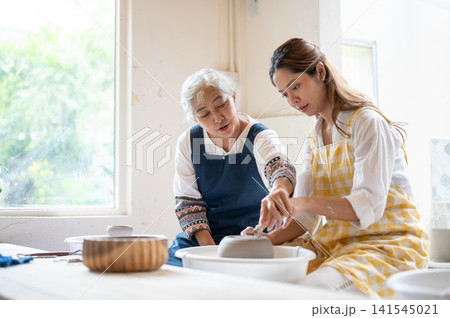 Senior Asian Instructor Guiding Student Hand to Trim Clay Bowl in pottery workshop studio. Senior Asian Instructor Guiding Student Hand to Trim Clay Bowl in pottery workshop studio. 141545021