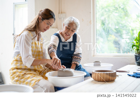 Senior Asian Woman Helping Young Student Trim Clay Bowl Base in pottery workshop studio. 141545022