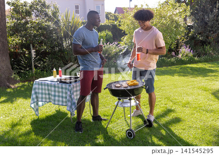Diverse male friends grilling corn, burgers on charcoal grill in backyard holding drinks Diverse male friends grilling corn, burgers on charcoal grill in backyard holding drinks 141545295