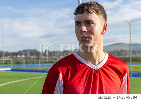 Male athlete in red jersey standing on turf field gazing left toward goal posts, copy space 141545394