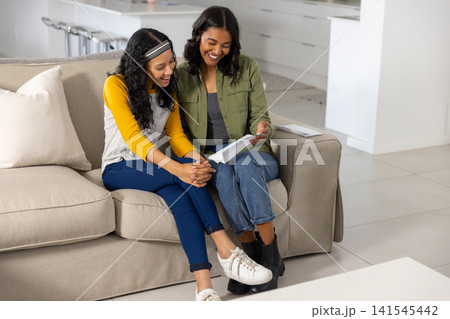 Diverse mom and teenage daughter reading letter from envelope while smiling on sofa at home 141545442