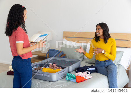 Diverse mom and teenage daughter packing grey suitcase with guidebook on bed in bedroom 141545461