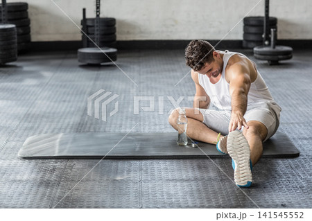 Man in 20s in tank stretching on gym mat, reaching toes to bottle, plates, copy space 141545552