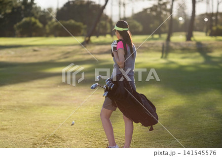 Female golfer standing on sunlit fairway carrying golf bag, wearing neon visor, golf ball nearby Female golfer standing on sunlit fairway carrying golf bag, wearing neon visor, golf ball nearby 141545576