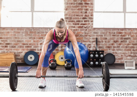 Adult female athlete wearing pink tank top and blue leggings, bending over loaded barbell at gym Adult female athlete wearing pink tank top and blue leggings, bending over loaded barbell at gym 141545598