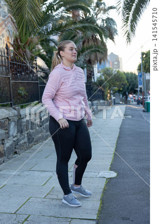 Woman standing on sidewalk beside low wall and palm trees, wearing pink hoodie and black leggings Woman standing on sidewalk beside low wall and palm trees, wearing pink hoodie and black leggings 141545710