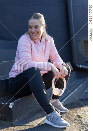 Woman in twenties smiling sitting on concrete steps at street holding headphones wearing pink coat Woman in twenties smiling sitting on concrete steps at street holding headphones wearing pink coat 141545716