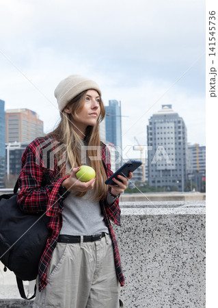 Adult female standing at concrete waterfront railing, holding phone and green apple, wearing beanie Adult female standing at concrete waterfront railing, holding phone and green apple, wearing beanie 141545736