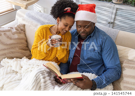 African American couple sitting on sofa at home, man wearing Santa hat, woman holding gray mug African American couple sitting on sofa at home, man wearing Santa hat, woman holding gray mug 141545744