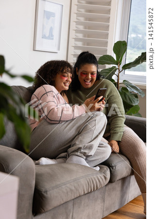 African American women sitting on grey sofa sharing phone, wearing pink patches and green knits African American women sitting on grey sofa sharing phone, wearing pink patches and green knits 141545872