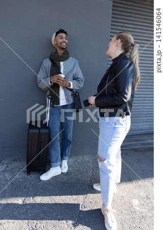 Diverse couple chatting on sidewalk near metal shutter, man wearing beanie holding suitcase, coffee Diverse couple chatting on sidewalk near metal shutter, man wearing beanie holding suitcase, coffee 141546064