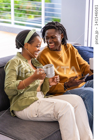 African American couple laughing holding mugs woman wearing headband man holding phone on gray sofa 141546081