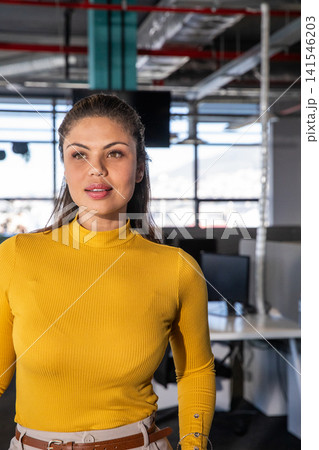 Woman standing, glancing right of camera in office with windows and monitors, wearing mustard top 141546203