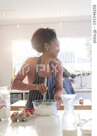 Diverse friends whisking batter in home kitchen using glass bowl and whisk, wearing striped apron 141546208