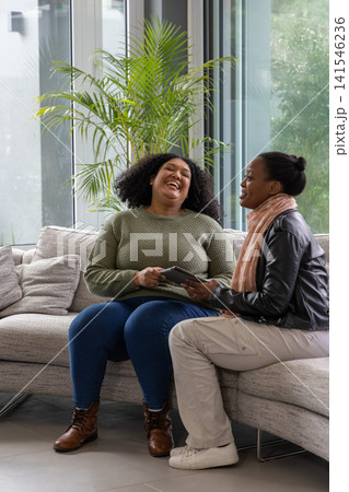 African American women laughing and talking while sharing tablet on sectional sofa by glass windows African American women laughing and talking while sharing tablet on sectional sofa by glass windows 141546236