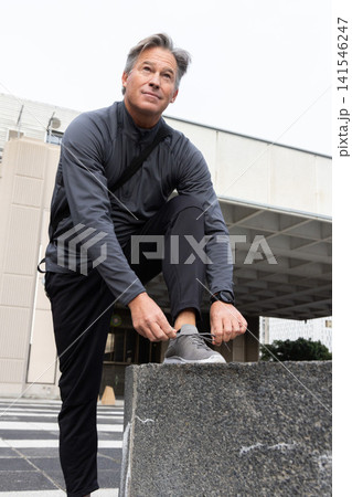 Middle-aged man in athletic clothing kneeling, tying gray running shoes on concrete block at plaza Middle-aged man in athletic clothing kneeling, tying gray running shoes on concrete block at plaza 141546247