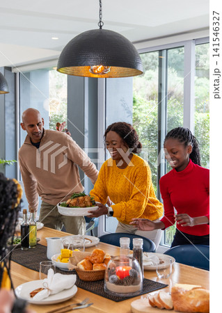 African American family carrying and serving roasted chicken at dining table with napkins, rolls 141546327