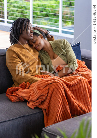 African American couple cuddling on gray sofa by deck under orange throw, wearing mustard sweater African American couple cuddling on gray sofa by deck under orange throw, wearing mustard sweater 141546344