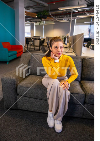 Woman sitting on gray sofa in open-plan office lounge wearing mustard top and checking smartwatch Woman sitting on gray sofa in open-plan office lounge wearing mustard top and checking smartwatch 141546410