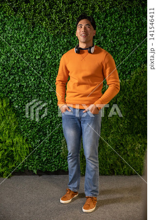Asian male mid 20s standing on carpet before green foliage wall in orange sweatshirt and headphones 141546411