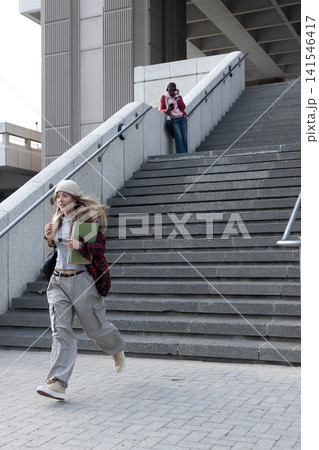 Diverse students running across urban plaza carrying notebooks and folder, one using smartphone Diverse students running across urban plaza carrying notebooks and folder, one using smartphone 141546417