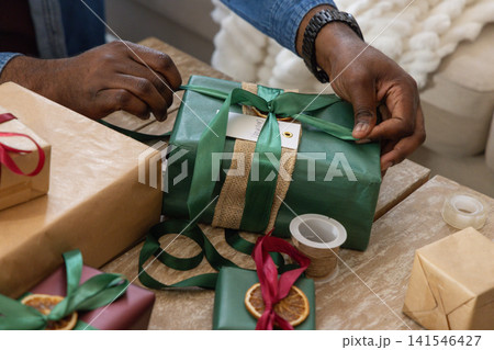 African American adult male denim sleeve hands tying green ribbon on green gift on coffee table 141546427