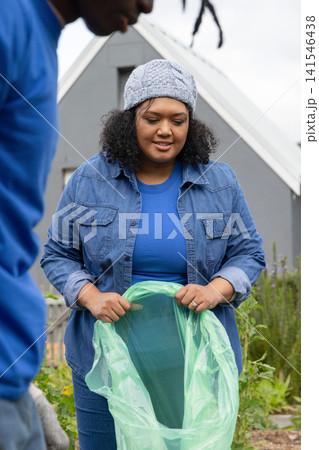 African American volunteers holding sheer green plastic bag in garden by grey home, with knit hat 141546438